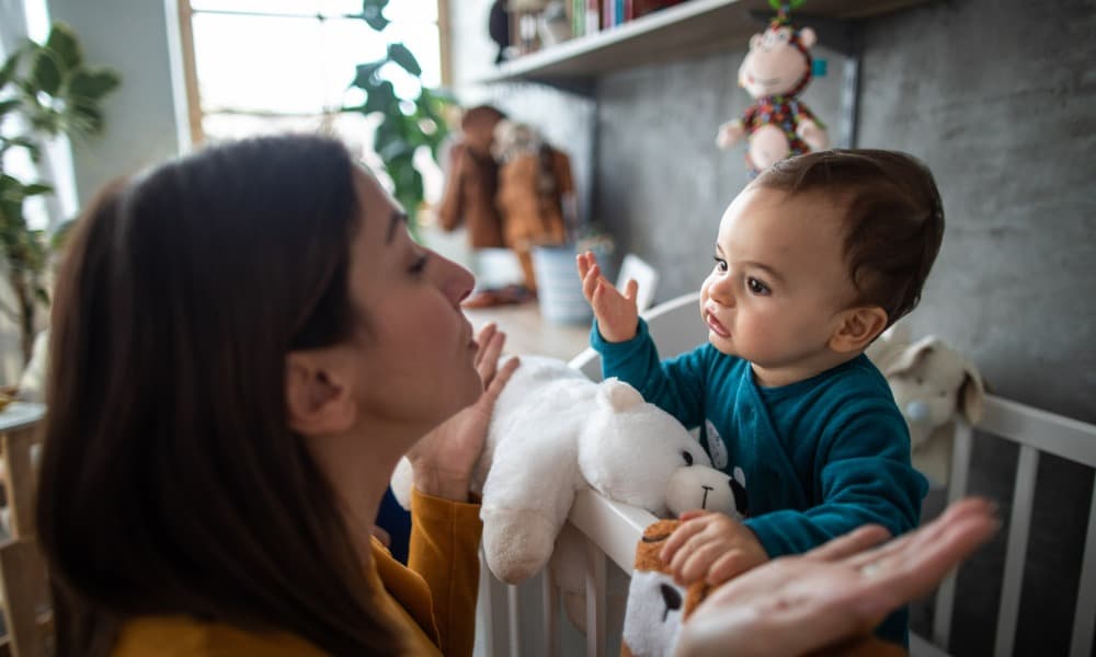 A mother talks to her baby who is standing in a crib 