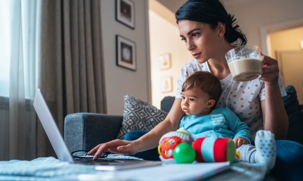 A mom holding a baby and a cup of coffee while working  from home 